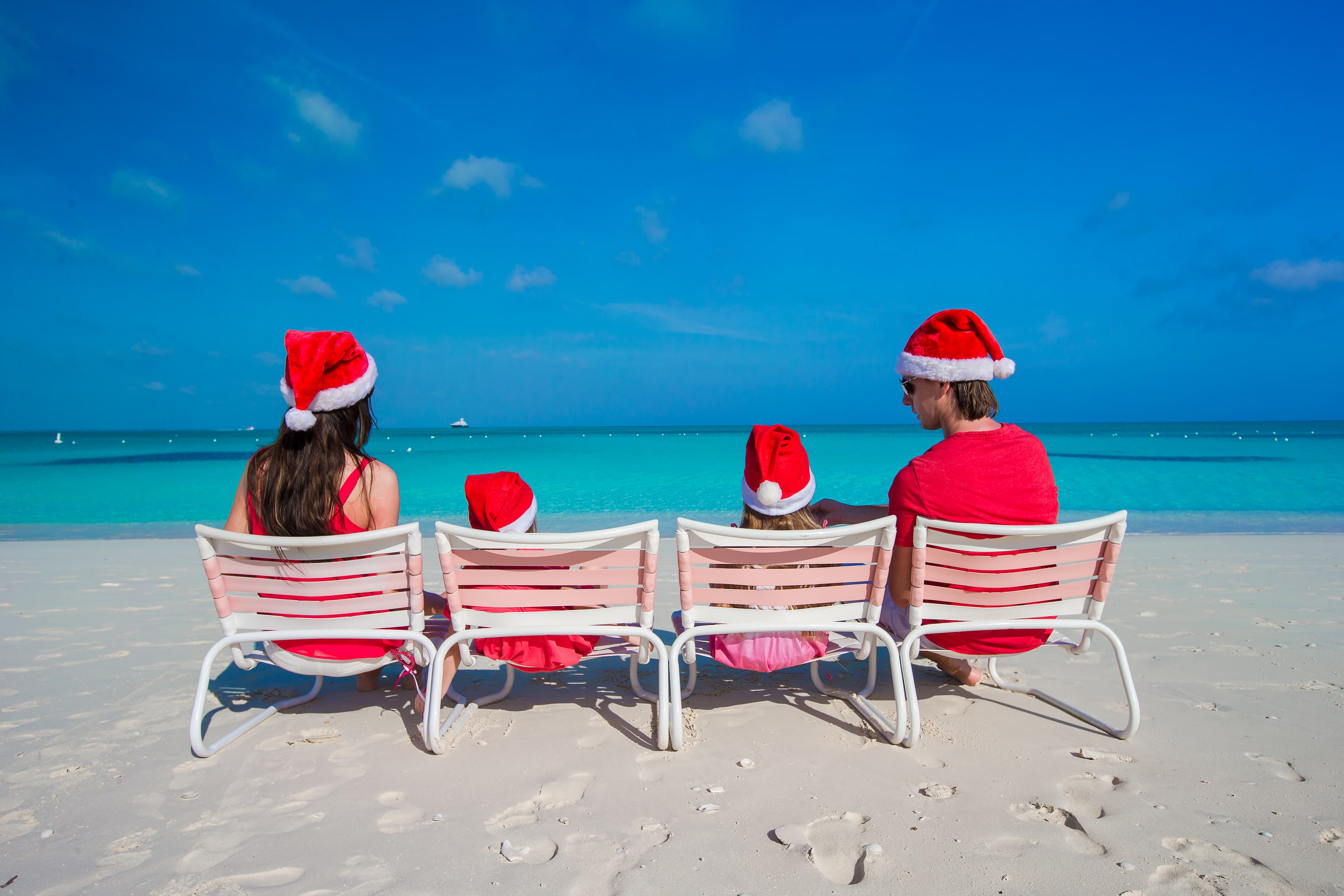 A family of four sits in beach chairs on the sand at the ocean while wearing bright red Santa hats