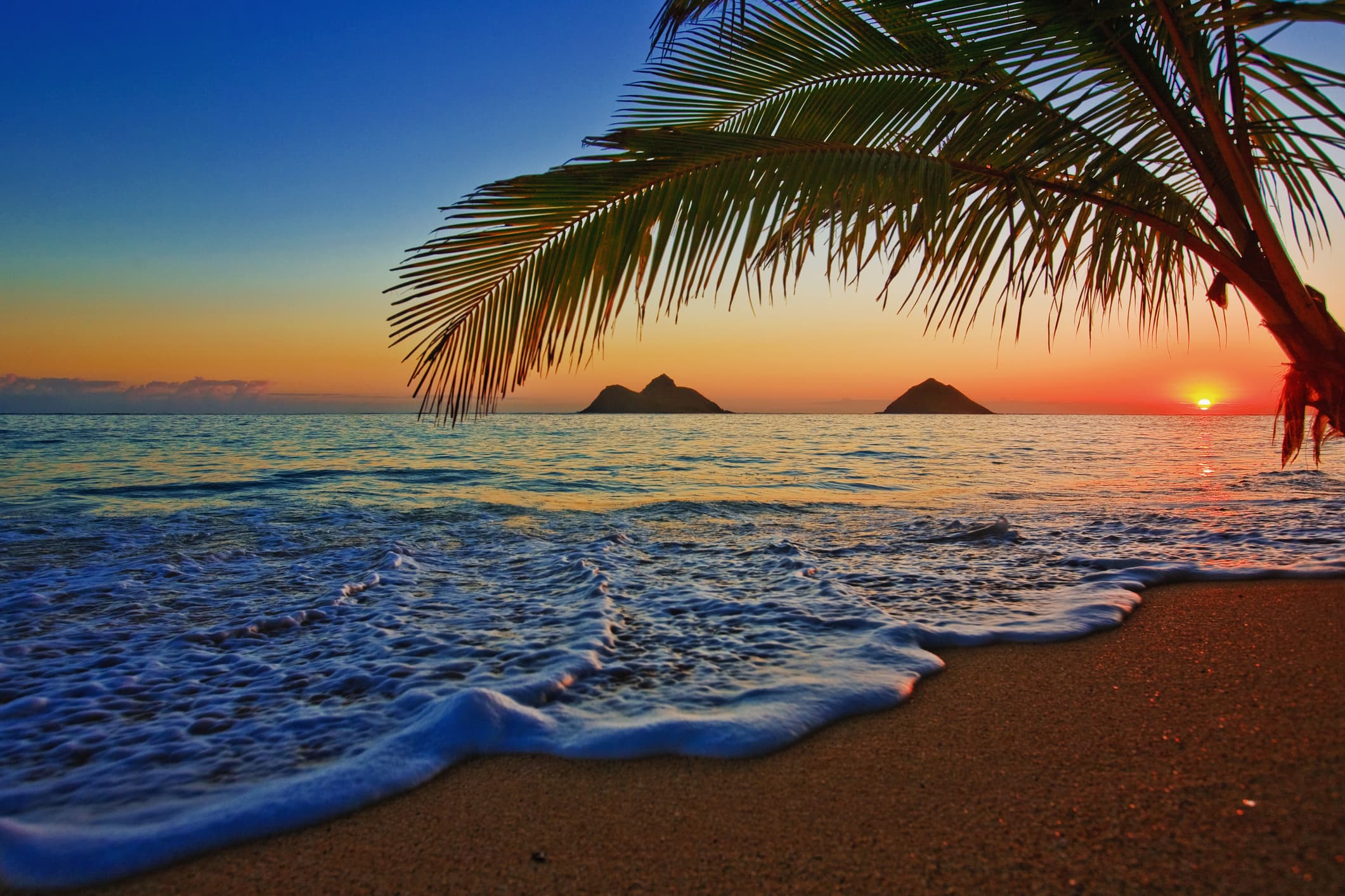 A waterside view of Lanikai Beach, Oahu, Hawaii, with gentle surf, palm trees, and sunset.