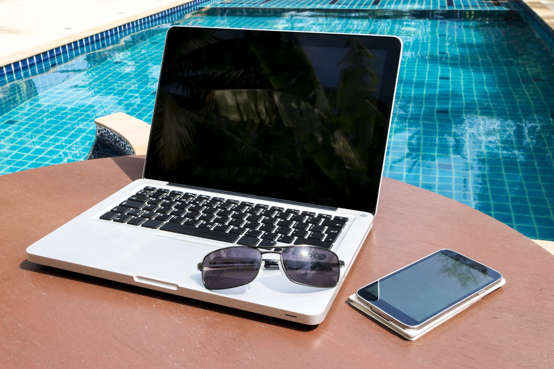 A view of a swimming pool with a laptop, smartphone, and sunglasses in the foreground.